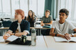 © Maskot - Non-binary person sitting by male friend at desk in classroom