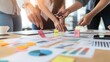 © LOGYARTISME99 - Closeup of a team of four hands collaborating on a project by adding sticky notes to a table covered with charts and graphs.