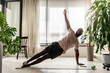 © lordn - Young African-American man doing a side plank exercise on a yoga mat at home, practicing strength and balance in a bright living room with indoor plants.