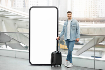 A young man stands beside a large blank display screen at a busy transportation hub, holding a suitcase. His casual attire contrasts with the sleek architecture around him.