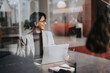 © Maskot - Smiling businesswoman sitting with laptop at desk seen through glass
