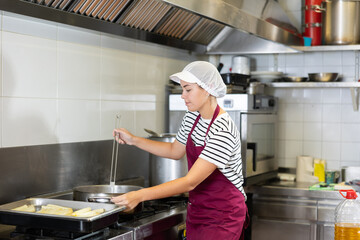 Skilled young woman in maroon apron and hairnet boiling asparagus in professional kitchen, preparing food for sale in deli section of local grocery store..