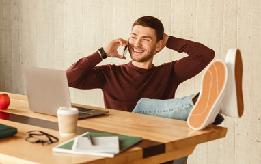 Poster - Successful Young Entrepreneur Talking On Cellphone Sitting At Laptop With Legs On Desk In Modern Office. Selective Focus
