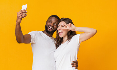 Wall Mural - Cheerful interracial couple taking selfie with smartphone, having fun together over yellow background. Playful girl showing peace gesture, empty space