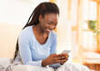 © Prostock-studio - Joyful Afro Lady Using Cellphone Playing Mobile Games Sitting In Bed At Home. Selective Focus