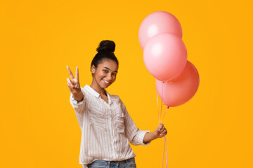 Portrait of laughing afro girl with pink air balloons showing peace sign while posing on yellow background in studio, copy space