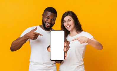 Poster - Great app.Happy multiracial couple holding and pointing at smartphone with black screen for mockup, standing together over yellow background