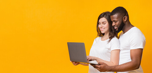 Wall Mural - Online Shopping Website. Portrait Of Happy Interracial Couple Using Laptop, Looking At Computer Screen Together, Yellow Background, Panorama