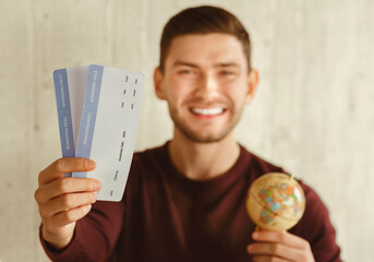 Poster - Vacation. Smiling Traveler Guy Showing Flight Tickets Holding World Globe Ready For Journey Sitting Indoor. Selective Focus, Shallow Depth