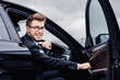 © Prostock-studio - Smiling manager in glasses sitting in luxury car, getting in or out his auto, keeping door open, looking at camera