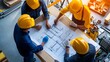 © Jiraphat - A group of construction workers in yellow hard hats collaborate over blueprints on a table, discussing plans for a project.