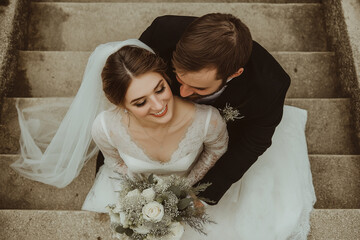 Wall Mural - A bride and groom posing for the camera, a top-view shot, wearing a white wedding dress with a veil and holding a bouquet. She is sitting on concrete stairs with her husband standing behind her