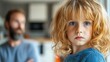 © Anna - A young child with bright blue eyes and curly hair looks contemplatively at the camera, with an adult blurred in the background. Soft light fills the cozy indoor space
