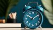 © Alfazet Chronicles - Close-up of a blue analog alarm clock on a desk with blurred background including books and plant, depicting time and organization.