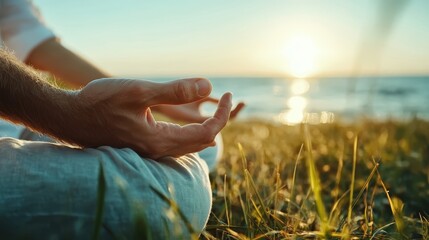 Close-up of hands in a meditative gesture while sitting on the grass near the seaside during a warm sunset, symbolizing inner tranquility and peaceful reflection.