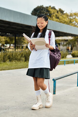 A cheerful young Asian girl in a stylish outfit reads a book outdoors, embracing youth culture and diversity.