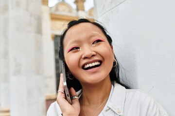 A cheerful young woman in a stylish outfit chats excitedly on her phone outside a university.