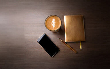 Wall Mural - A flat lay showcasing a cup of coffee with latte art, a smartphone, a pen, and a gold notebook on a wooden table.