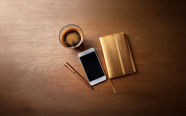 Wall Mural - A wooden table with a glass of coffee, a smartphone, a pen, and a gold notebook.