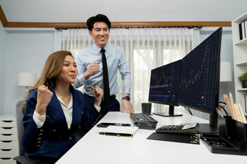 Successful smiling Asian business partners raising fist up together in profitable project job in stock exchange database on pc wearing formal suit at modern home office at side view image. Infobahn.