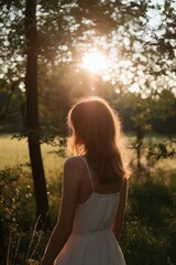 Wall Mural - A woman stands in the forest, gazing at the setting sun as light filters through the trees.