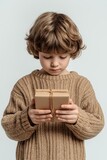 Young child with soft curls holding a small wrapped gift box, wearing a cozy beige sweater and looking down with a thoughtful expression, isolated on a light background