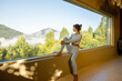 © rh2010 - A woman in a light-colored yoga outfit sits on a window ledge, gazing out at the misty mountains and lush greenery outside. The serene indoor setting enhances the peaceful moment of reflection