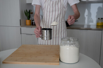 man with apron sifts flour through a sieve in the domestic kitchen