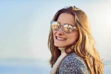 Happy woman, sunglasses and smile with fashion or accessory by beach or ocean coast in nature. Young, female person or tourist with travel in joy for style, summer season or holiday weekend by sea