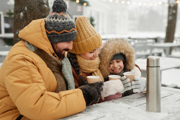 Wall Mural - Portrait of modern young family drinking hot cocoa outdoors in winter sitting at cafe table together and smiling happily