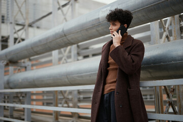 Fashionable man in brown coat talking on cell phone in city, standing in front of metal railing on building side