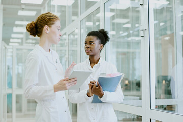 Two female scientists in laboratory coats discussing research data in modern laboratory, showing deep focus and engagement in their work