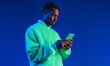 © Krisartist94 - Young man wearing a bright green hoodie interacts with a smartphone against a vibrant blue background during the evening