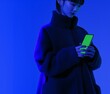 © Krisartist94 - Young woman in a cozy coat interacting with a smartphone under vibrant blue and green lighting in an indoor space