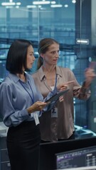 Poster - Vertical Screen: Two Female Data Center Engineers, an Asian and Caucasian Women, Collaborate Using a Tablet in a Glass Office Next to a Futuristic Server Room, Analyzing Software Data on Screen