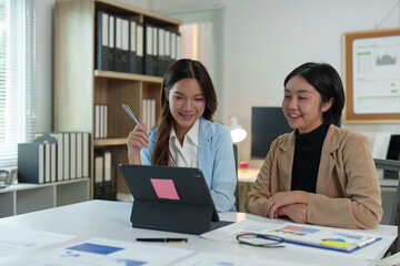 Two young businesswomen pointing to plans, statistics, and data graphs and explaining the conditions of their partnership, happily acknowledging their turnover and income during negotiations.