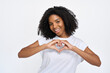 © insta_photos - Happy young African American woman wearing t-shirt showing heart sign hand gesture standing isolated at white wall background. Gen z female model expressing concept of love.