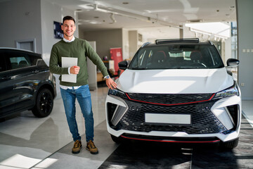 A man stands on the right side of a white car in a dealership, holding papers in his right hand. He is cheerful, touches the vehicle with his left hand, and looks at the camera with excitement.