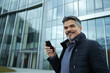 © Impressionstock - Happy businessman in casual jacket holding smartphone outside modern glass building in daylight | Corporate success and positive communication concept with business executive smiling confidently.
