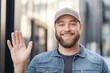 © Vasili - Bearded man in a baseball cap waving and smiling while standing on a city street. The image captures a warm and friendly gesture in a bright outdoor setting.