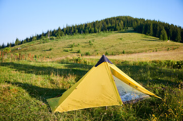 Wall Mural - Bright yellow tent on lush grassy meadow under clear blue sky. On background gently sloping hill covered with scattered trees and shrubs, creating serene and picturesque camping scene.