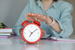 © Pawel - Selective focus of an alarm clock on a table next to a person using a notepad to write at home, a time management concept
