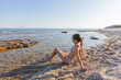 © mitarart - A young girl in a swimsuit sitting by the clear beach waters, enjoying a summer day.