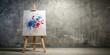 © chaiwat - Aerial view of large white canvas with red and blue paint splatters on gray wall background with wooden easel