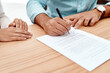 © peopleimages.com - Person, hands and writing with signature on contract for insurance, application or legal agreement on wooden table. Closeup, people or signing with paper for deal, policy or employment form on desk