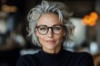 © Odin AI - A confident woman with gray hair and stylish glasses smiles warmly, exuding elegance and poise, set against a softly blurred background with warm lighting.