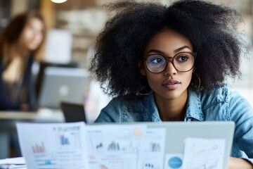 Poster - A woman with curly hair and glasses keenly examining papers with graphs and charts, using a laptop in a modern workspace with blurred background.