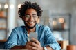 © zhuan - Happy young entrepreneur using smartphone at desk, smiling man with glasses and blue shirt holding phone, office workspace