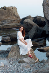 Poster - Serene woman in white dress sitting on rock by water with rocky landscape in background