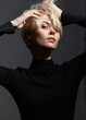 © SHOTPRIME STUDIO - Stylish young woman with short blonde hair, wearing a black turtleneck, striking a confident pose against a dark background. Modern fashion and beauty concept.